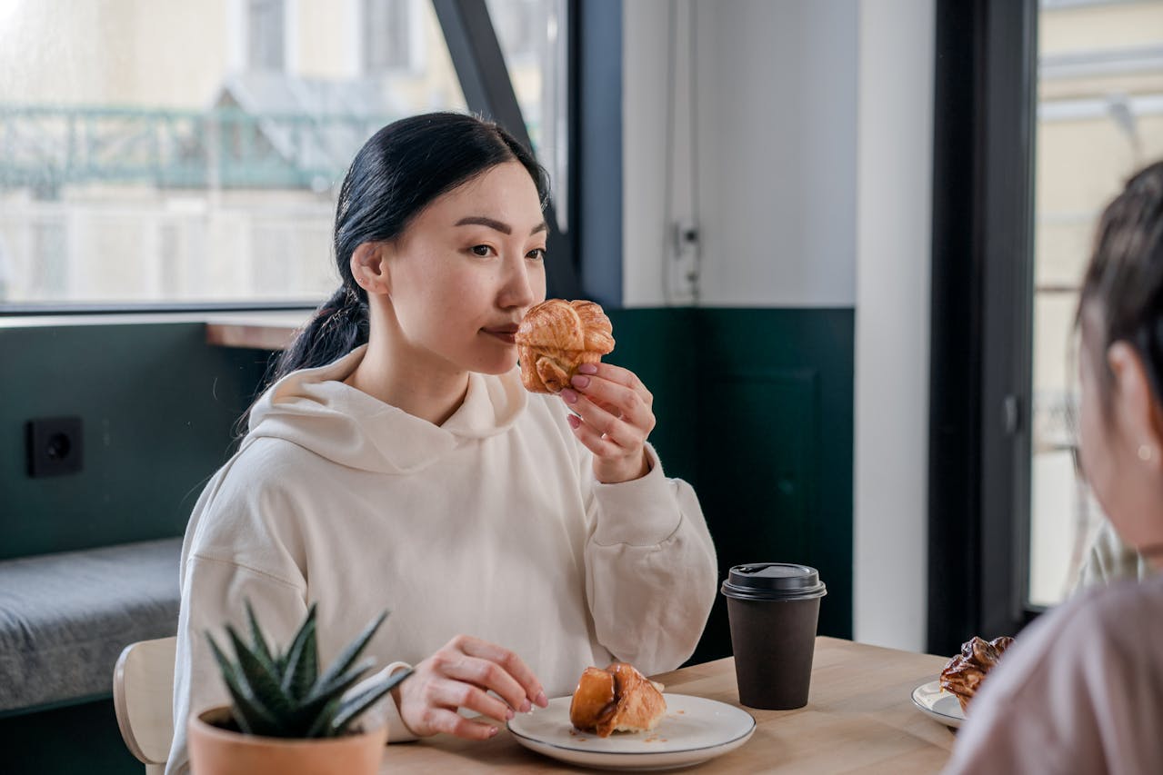 An asian woman in a cream colored hoodie sitting in a modern cafe smells a muffin